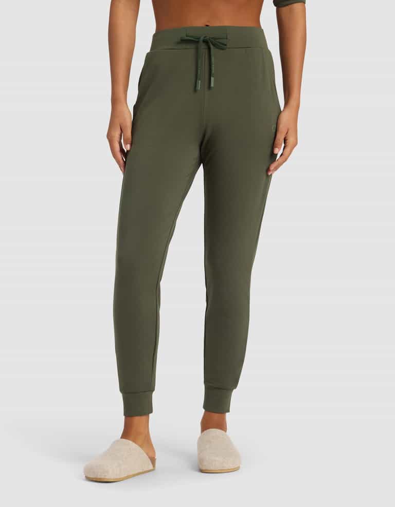 A person wears Cozy Earth's Women's Brushed Bamboo Jogger Pant in olive, featuring a drawstring waist and side pockets, paired with light beige slippers. Only the lower torso and legs are visible against a plain background.|Color:Olive