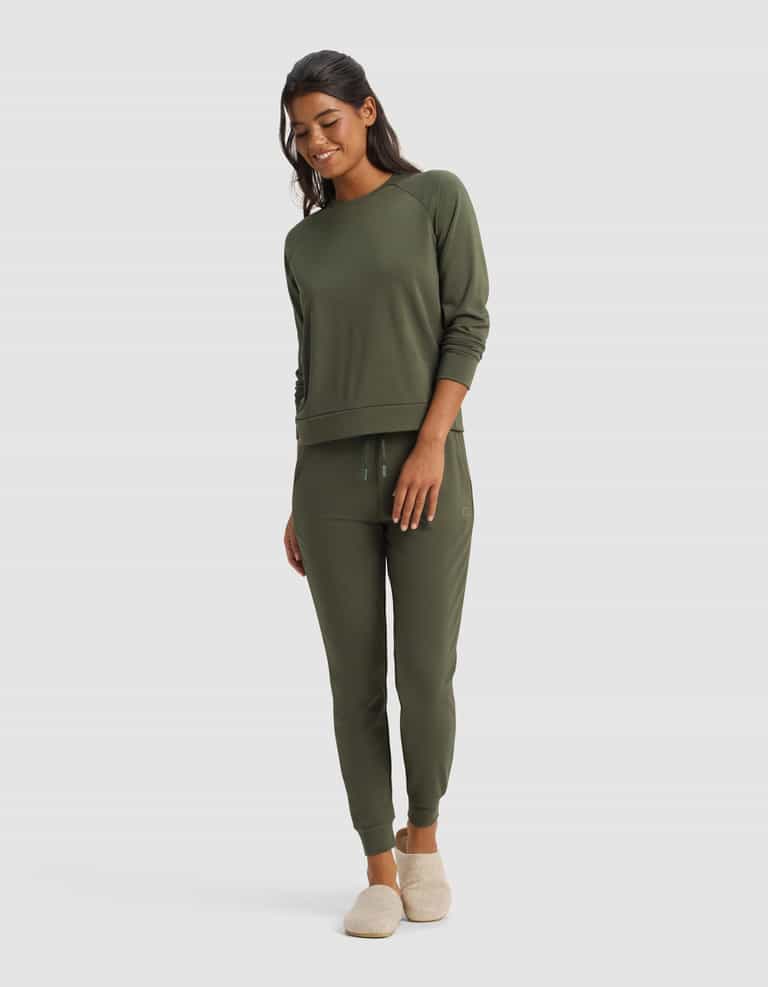 A woman smiles in Cozy Earth's Women's Brushed Bamboo Jogger Pant, paired with a matching olive green sweatshirt and beige slippers, set against a plain light background.|Color:Olive