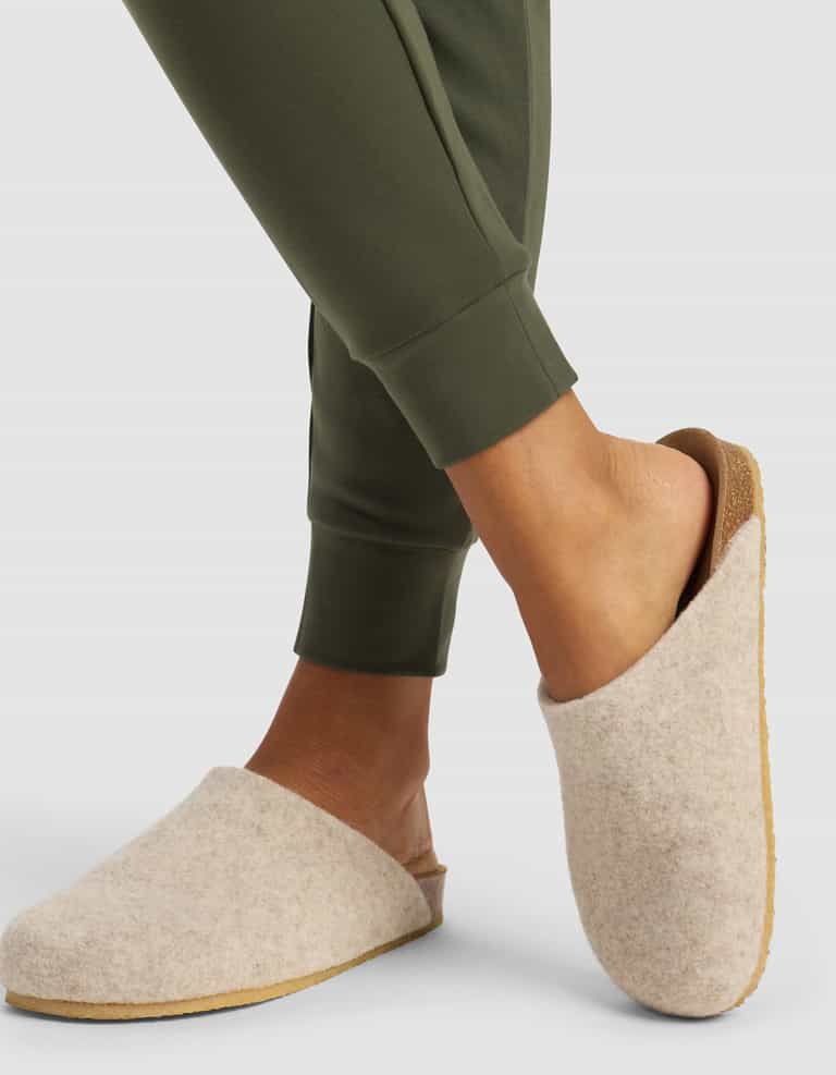 A woman models the Cozy Earth Women's Brushed Bamboo Jogger Pant in olive green, paired with light beige wool slippers, standing with one foot crossed over the other against a plain background.|Color:Olive