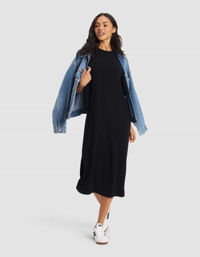 A woman poses against a plain light background wearing Cozy Earth's Women's Brushed Bamboo Midi Dress in black, paired with white sneakers and a blue denim jacket draped over her shoulders.|Color:Black