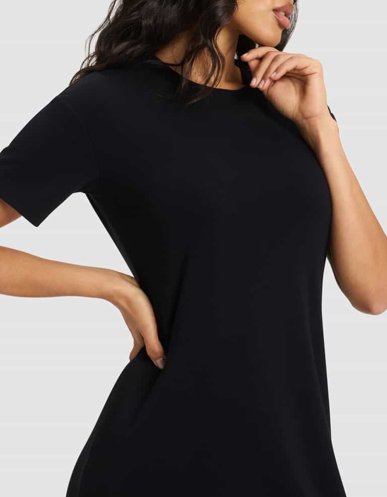 A woman models the Cozy Earth Women's Brushed Bamboo Midi Dress, a short-sleeved black style, posing with one hand on her hip and the other near her face against a plain light gray background. Her face is partially visible.|Color:Black