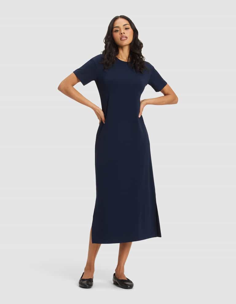 A woman with long dark hair stands against a plain background wearing Cozy Earth's Women's Brushed Bamboo Midi Dress in navy, featuring short sleeves, an ankle-length hem with side slit, and paired with black flats. Her hands rest on her hips.|Color:Navy
