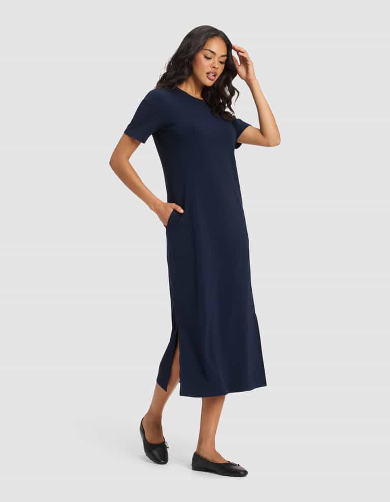 A woman wearing the Cozy Earth Women's Brushed Bamboo Midi Dress in navy blue with short sleeves and side slits, paired with black flats, stands against a light gray background, one hand in her pocket and the other touching her hair.|Color:Navy