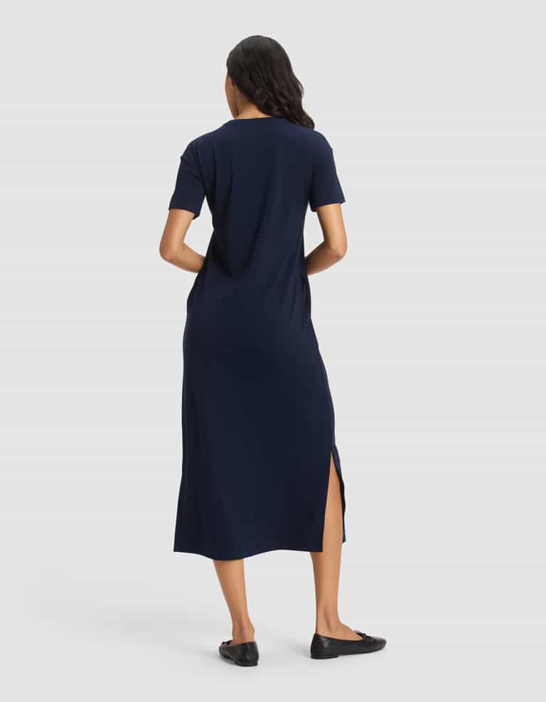 A woman with loose waves stands facing away on a plain white background, hands in her pockets, wearing Cozy Earth's Women's Brushed Bamboo Midi Dress in navy blue with short sleeves and side slit, paired with black flats.|Color:Navy