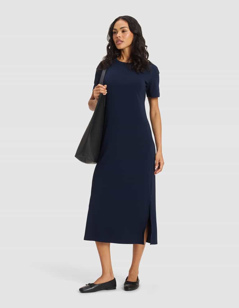 A woman wears the Cozy Earth Women's Brushed Bamboo Midi Dress in navy blue with short sleeves and a side slit, paired with black flats and a large black tote bag, standing against a plain light background.|Color:Navy