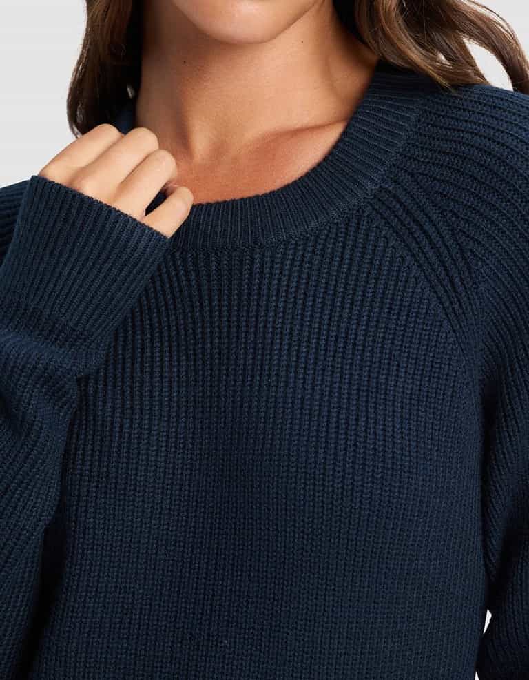 A woman wears the Cozy Earth Women's Classic Crewneck in dark blue, shown from shoulders to upper chest with her hand gently holding the collar. Her brown wavy hair is visible, and her face is partly shown.|Color:Eclipse