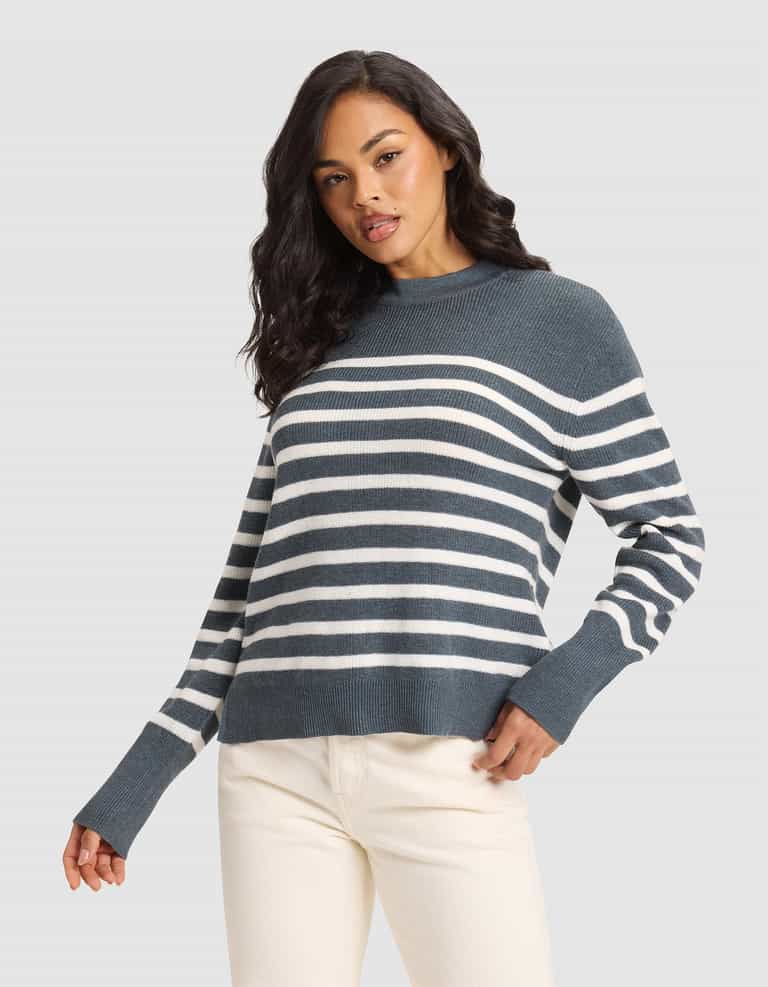 A woman with long dark hair models the Cozy Earth Women's Rowan Sweater—a blue and white striped knit—paired with off-white pants against a plain light gray background, one hand in her pocket and a relaxed expression. |Color:Dawn/Bone Stripe