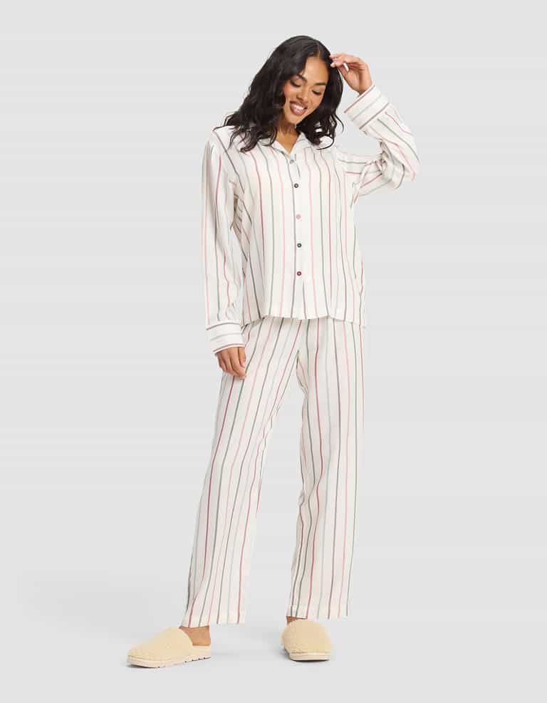 A woman smiles in a plain light gray setting, wearing the Cozy Earth Women's Soft Woven Long Sleeve Pajama Top in cream with vertical red and beige stripes, paired with beige slippers. |Color:Cottage Stripe