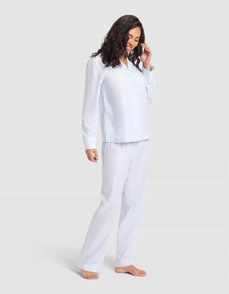 A woman with long dark hair, barefoot and smiling, models the Cozy Earth Women's Soft Woven Long Sleeve Pajama Set—light blue and white striped—against a plain light gray background. |Color:Spring Blue Stripe