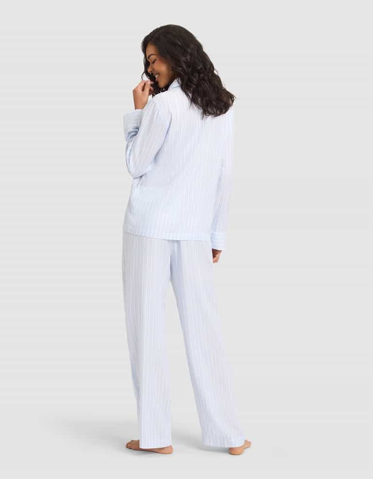 A woman stands barefoot, facing away in the Cozy Earth Women's Soft Woven Long Sleeve Pajama Set, light blue and white striped. She glances over her shoulder with a smile, hand near her mouth, against a plain light gray background. |Color:Spring Blue Stripe