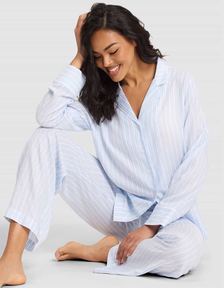 A woman with long dark hair sits barefoot on the floor, smiling and looking down, wearing the Cozy Earth Women's Soft Woven Long Sleeve Pajama Top in a light blue and white striped pattern. |Color:Spring Blue Stripe
