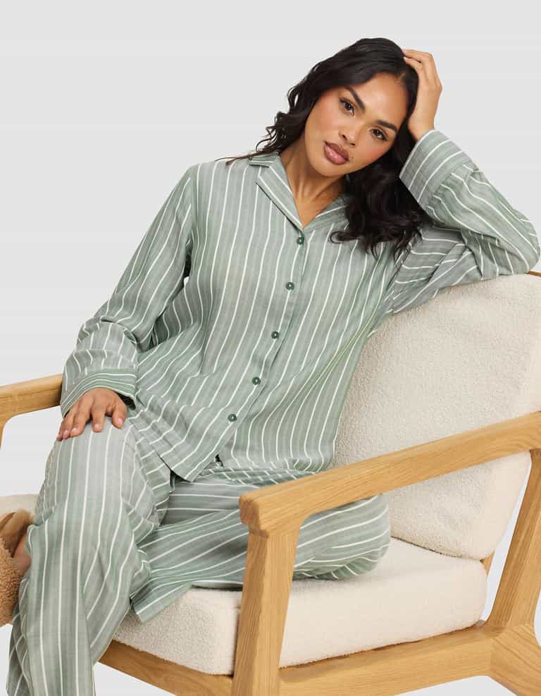 A woman in the Cozy Earth Women's Soft Woven Long Sleeve Pajama Set, featuring green and white stripes, sits comfortably on a light cushioned wooden chair and looks at the camera with a relaxed expression.|Color:Striped Jasper