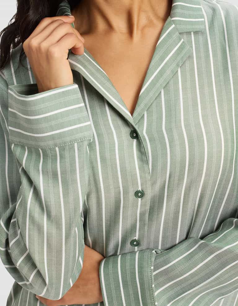 A woman wears the Cozy Earth Women's Soft Woven Long Sleeve Pajama Top, holding the collar with one hand and crossing her other arm over her body.|Color:Striped Jasper