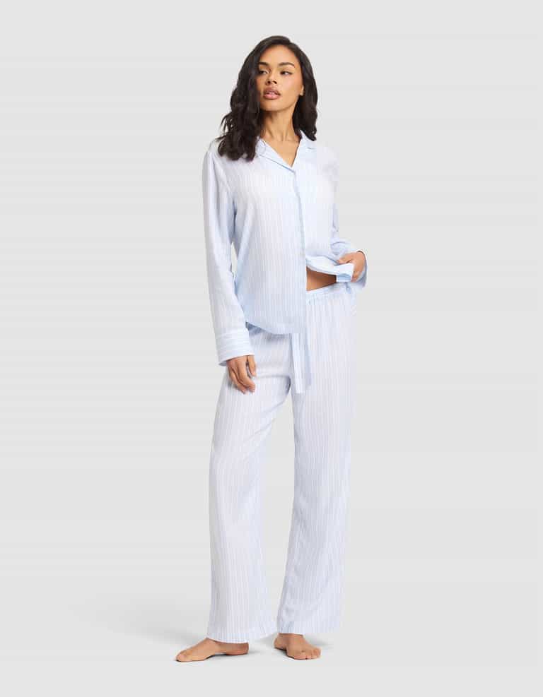 A woman stands barefoot against a light gray background wearing Cozy Earth Women's Soft Woven Pajama Pant in light blue and white stripes with a matching button-up shirt, her left hand in her pocket. |Color:Spring Blue Stripe