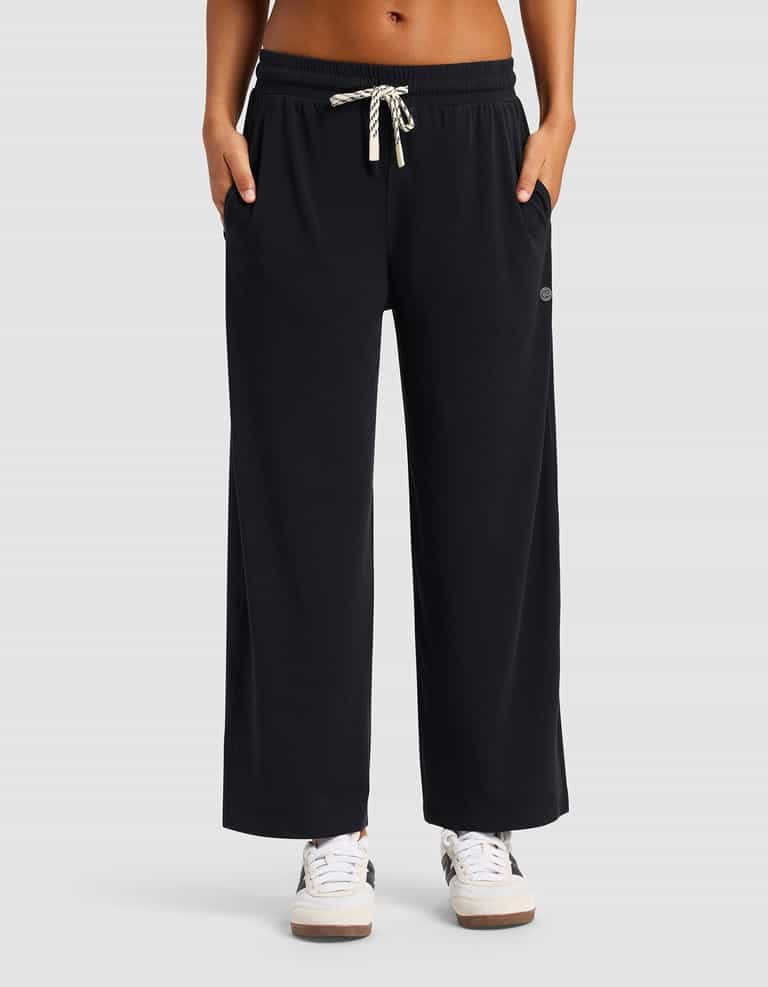 A woman wears Cozy Earth’s Women’s Studio Cropped Wide Leg Pant in black, featuring pockets and a drawstring waist, paired with white sneakers against a plain light gray background.|Color:Jet Black