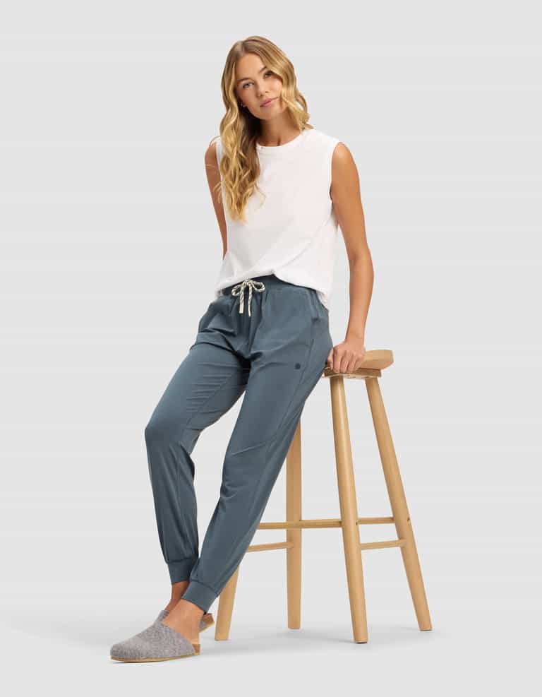 A woman with long blonde hair models Cozy Earth's Women's Studio Jogger in blue, paired with a white sleeveless top and gray slippers, sitting on a wooden stool against a plain light gray background.|Color:Dawn