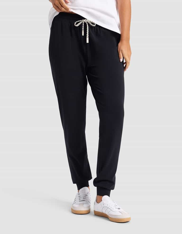 A person wearing Cozy Earth's Women's Studio Jogger in black with a white drawstring, a white T-shirt, and white sneakers stands against a plain light gray background.|Color:Jet Black