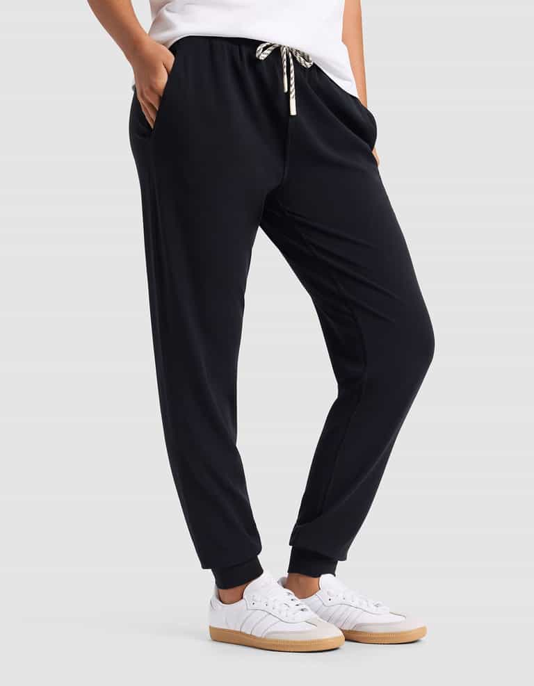 A woman wears Cozy Earth Women's Studio Jogger—black with a drawstring waist—paired with white sneakers and a white top, standing hands-in-pockets against a plain light gray background.|Color:Jet Black