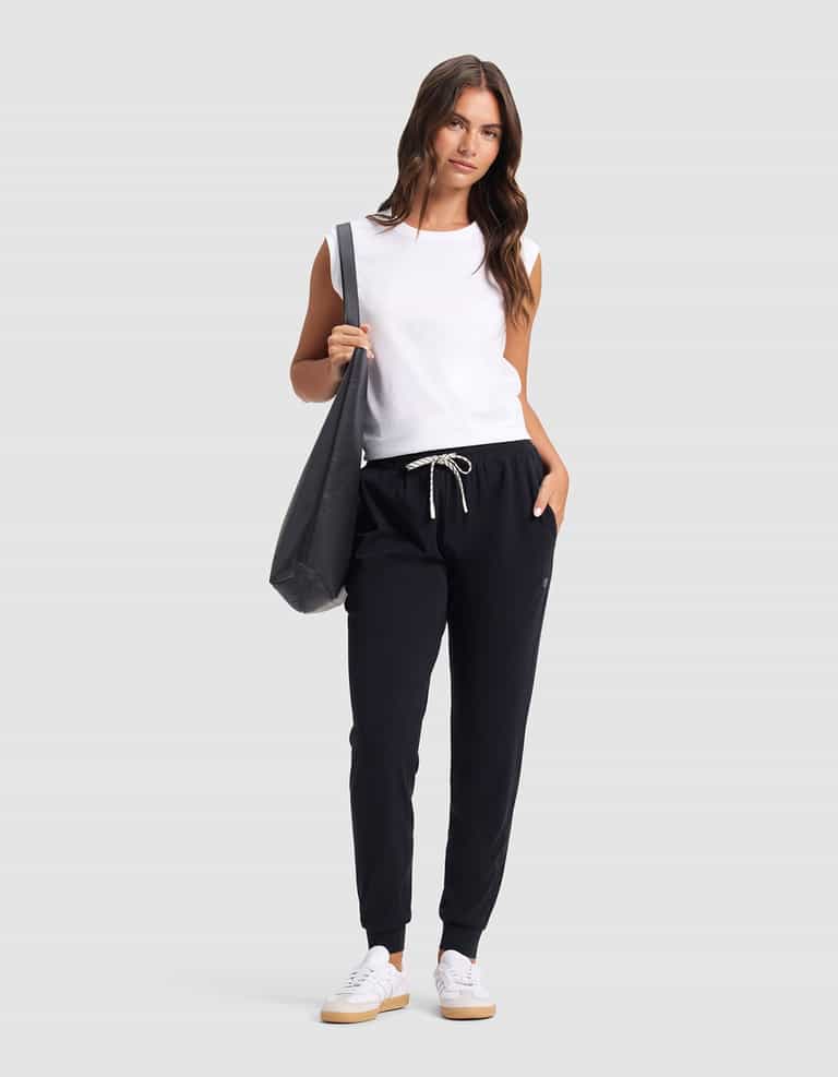 A woman poses against a plain background wearing Cozy Earth's Women's Studio Jogger in black with a drawstring, a sleeveless white top, white sneakers, and carrying a large black tote bag on her shoulder.|Color:Jet Black
