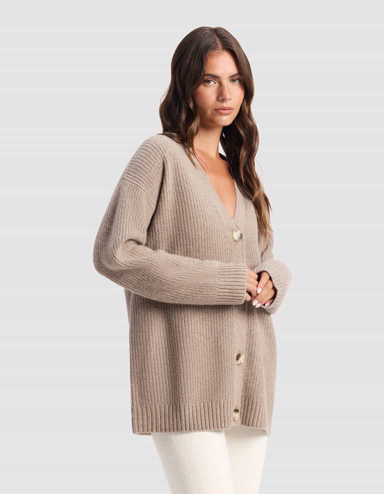 A woman with long brown hair wears Cozy Earth's Oversized Sunday Cashmere Cardigan in beige with large buttons and white pants, standing against a light gray background.|Color:Almond