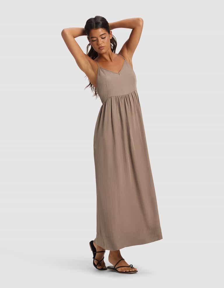 A woman models the Cozy Earth Women's Sunset Gathered Dress—a sleeveless, light brown maxi—paired with black strappy sandals, posing with arms raised against a plain light background.|Color:Ebony