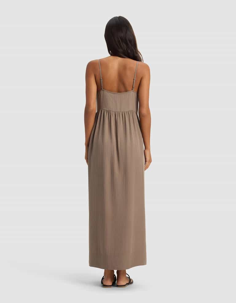 A woman with long dark hair, seen from behind, wears the Cozy Earth Women's Sunset Gathered Dress—sleeveless, ankle-length with thin straps—paired with flat sandals, standing against a plain light gray background.|Color:Ebony