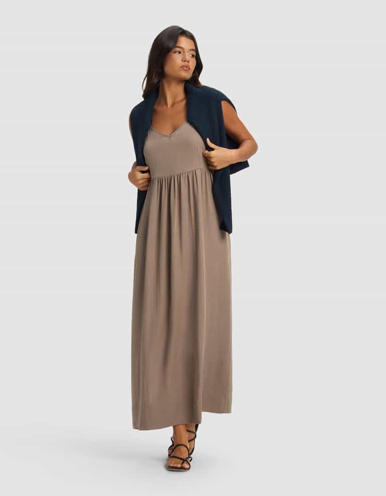 A woman models the Cozy Earth Women's Sunset Gathered Dress in taupe, styled with black sandals and a navy sweater over her shoulders, standing against a plain light gray background and looking slightly to the side.|Color:Ebony