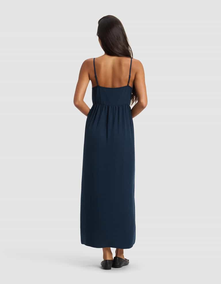 A woman with long dark hair stands facing away, wearing the Cozy Earth Women's Sunset Gathered Dress in navy blue with thin straps and black shoes, set against a plain white background.|Color:Eclipse