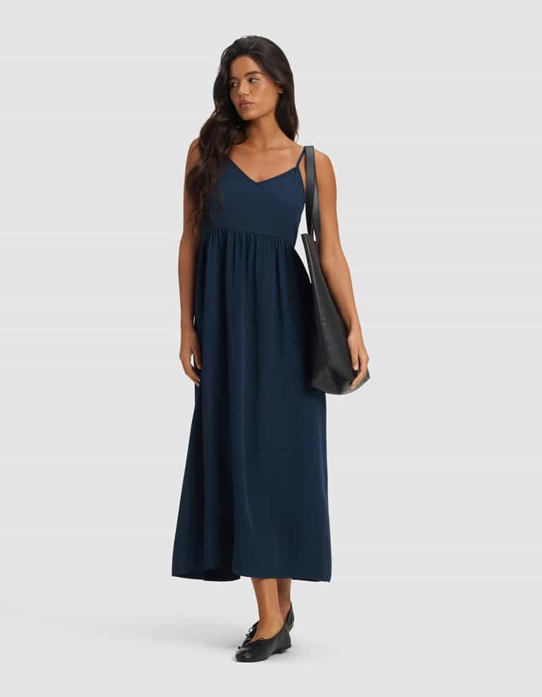 A woman in the Cozy Earth Women's Sunset Gathered Dress - Last Chance, a sleeveless navy maxi with a V-neckline, styled with black flats and a large black shoulder bag, stands against a plain light gray background.|Color:Eclipse