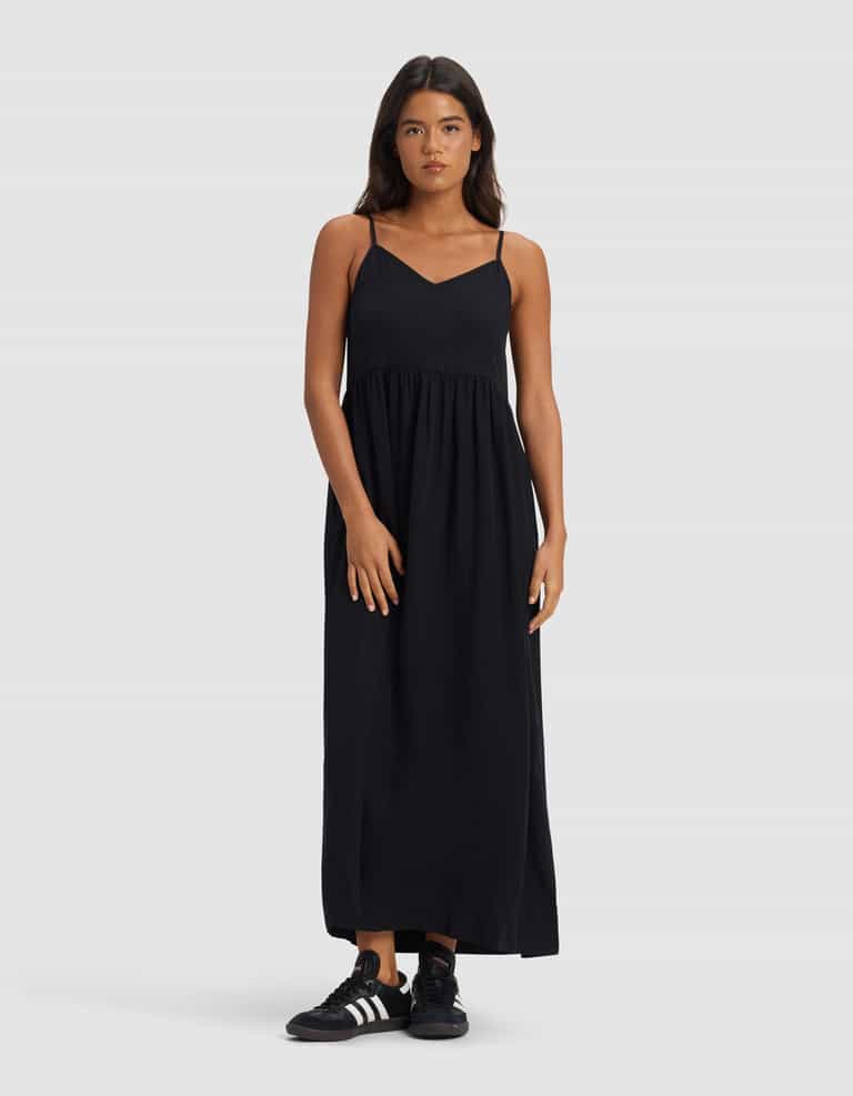 A woman faces forward in a Cozy Earth Women's Sunset Gathered Dress – Last Chance, an ankle-length, sleeveless black dress with thin straps, paired with black Adidas sneakers against a plain light gray background.|Color:Jet Black