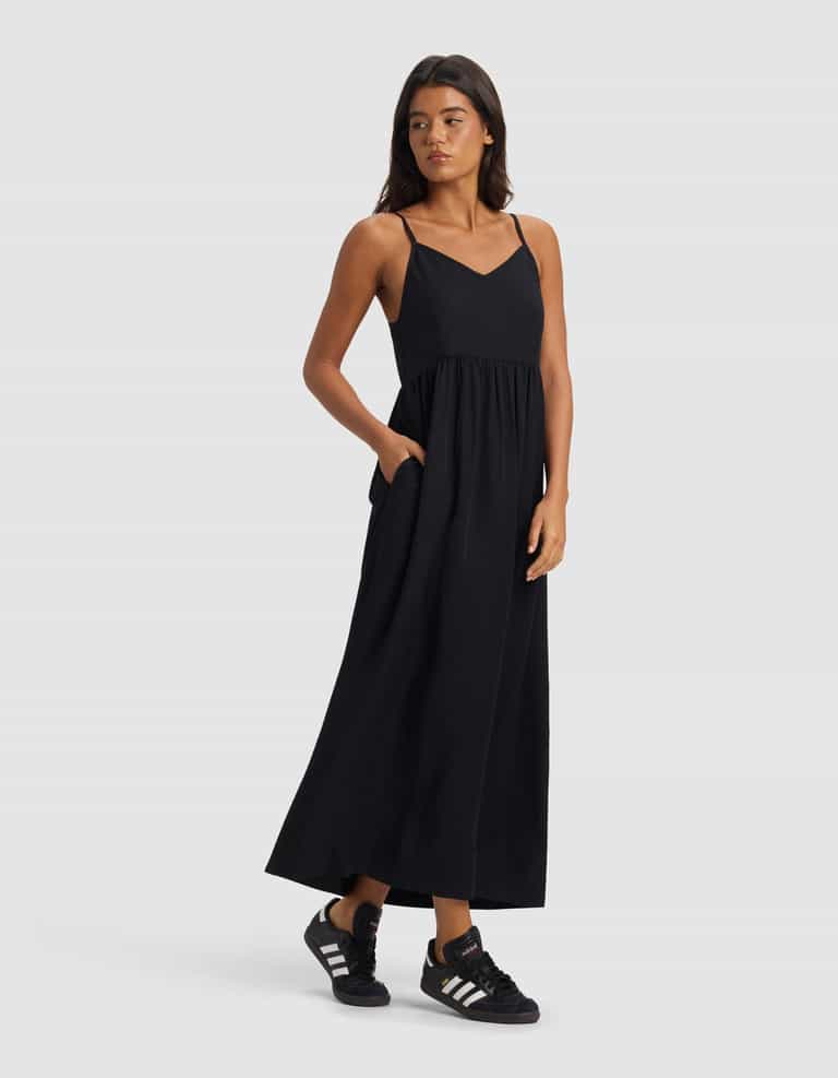A woman wearing the Cozy Earth Women's Sunset Gathered Dress - Last Chance stands against a plain light gray background, looking to her left with one hand in her pocket and pairing the dress with black Adidas sneakers.|Color:Jet Black