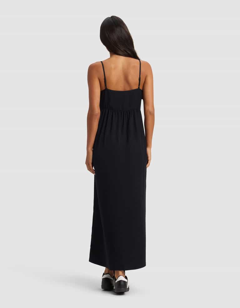 A woman with long dark hair stands facing away from the camera, wearing the Cozy Earth Women's Sunset Gathered Dress - Last Chance in black, styled with black shoes, set against a plain light background.|Color:Jet Black