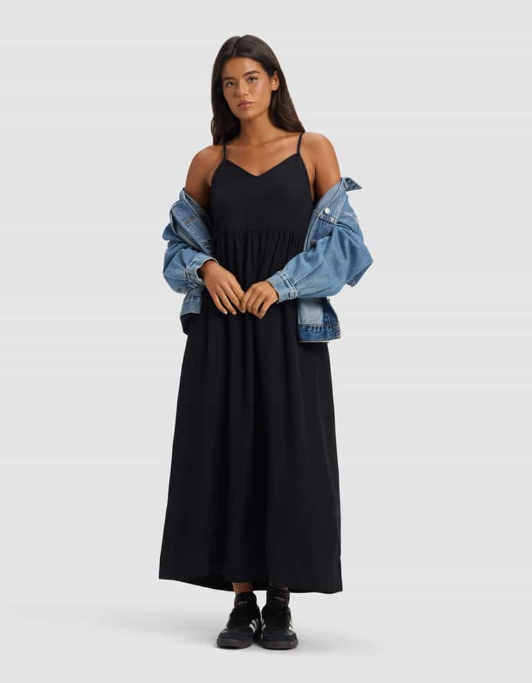 A woman with long dark hair and a neutral expression stands facing forward against a plain light grey background, wearing the Cozy Earth Women's Sunset Gathered Dress - Last Chance, with a blue denim jacket off her shoulders and black sneakers.|Color:Jet Black