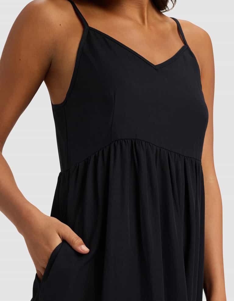 A model wears the Cozy Earth Women's Sunset Gathered Dress - Last Chance, a sleeveless black dress with thin straps and a V-neckline, shown from shoulders to mid-thigh, with one hand in the pocket.|Color:Jet Black