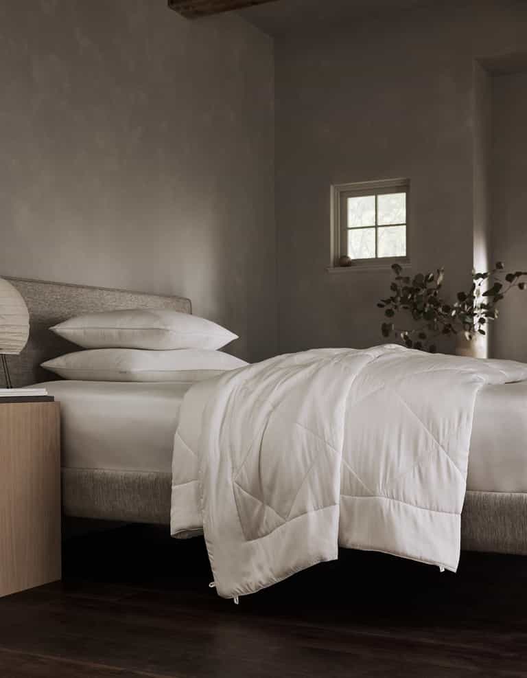A neatly made bed features the Cozy Earth Bamboo Viscose Comforter with white bedding and pillows in a softly lit, minimalist room. A small window, bedside table, and leafy plant are seen in the background.|Filling:Standard