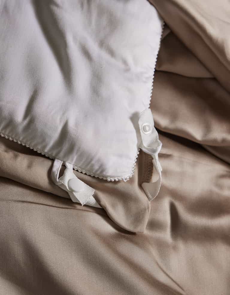 A close-up of Cozy Earth’s Bamboo Viscose Comforter with decorative trim and untied ribbon ties, resting on smooth, light beige sheets. The comforter's soft fabric appears slightly rumpled for a relaxed look.|Filling:Extra