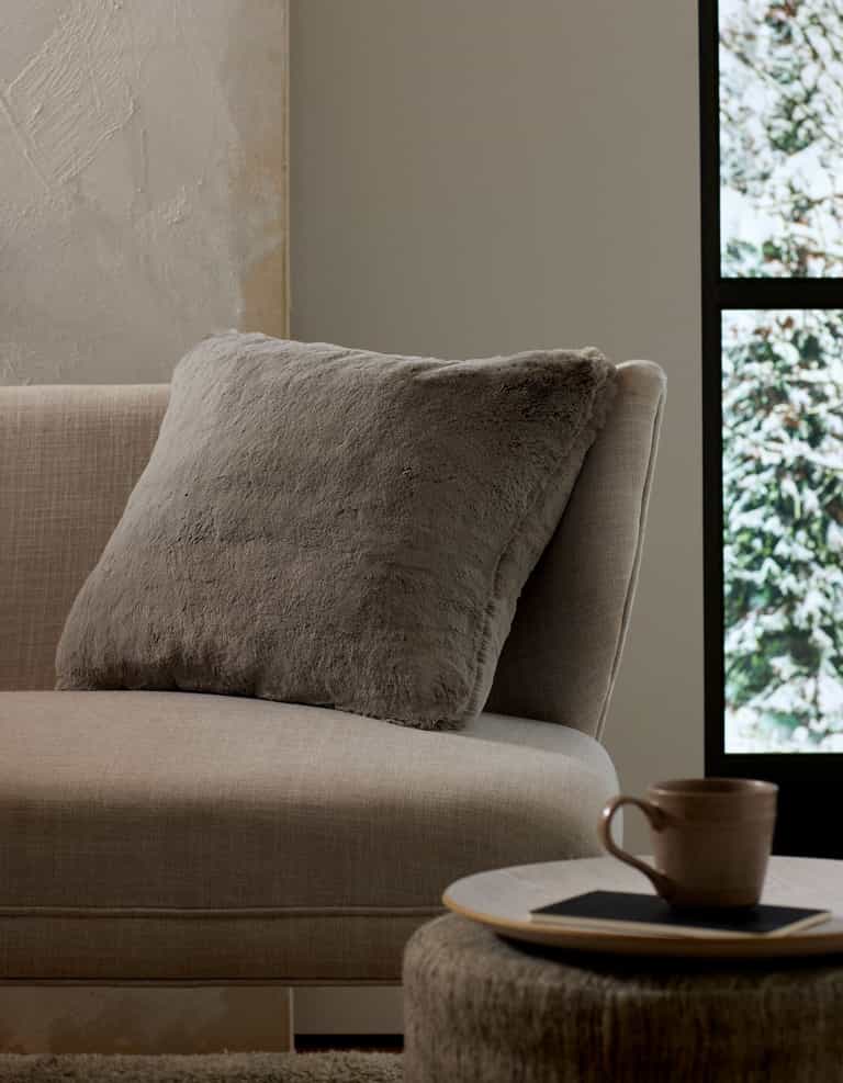 A cozy beige couch with a large, soft Cuddle Pillow by Cozy Earth rests beside a round table holding a ceramic mug and coaster. Sunlight pours through the window, revealing snowy trees outside.|Color:Dove Grey |Size:20" x 20"