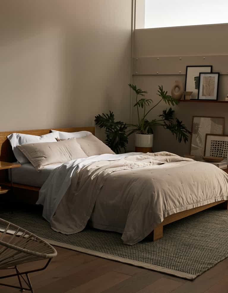 A cozy bedroom features a wooden bed frame with Cozy Earth's Soft-Wash Cotton Quilt in beige and white pillows. A large plant, framed artwork, and decor sit against the wall while soft natural light pours through a high window.|Color:Driftwood