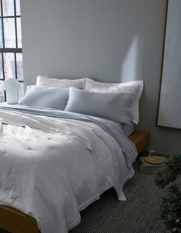 A neatly made bed with Cozy Earth's Soft-Wash Cotton Quilt in white and light gray sits by a window with soft natural light, next to a small round bedside table, lamp, and framed artwork. The room has calm, minimalist decor.|Color:White