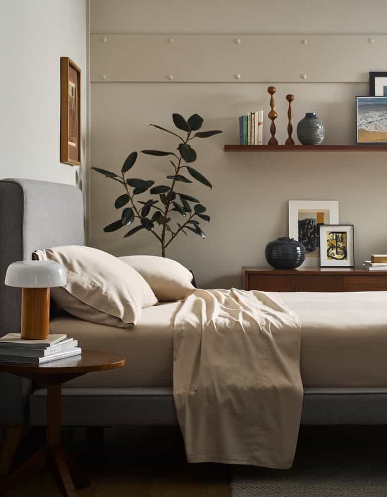 A bed made with Cozy Earth’s Soft-Wash Cotton Flat Sheet, a gray upholstered headboard, and a wooden nightstand holding a lamp and books. A leafy plant, artwork, and decor sit on a shelf against a light wall.|Color:Driftwood