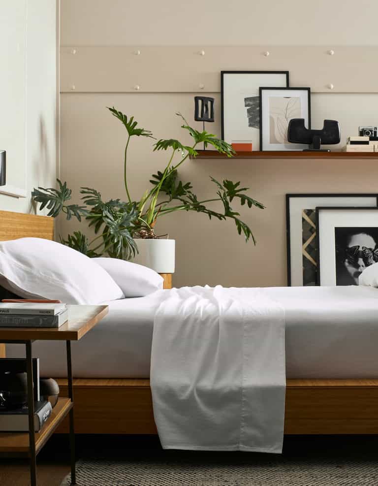 A modern bedroom features Cozy Earth's Soft-Wash Cotton Flat Sheet, a wooden bed with white bedding, a leafy plant in a white pot, framed art and decor on a shelf above the bed, and a side table with books.|Color:White