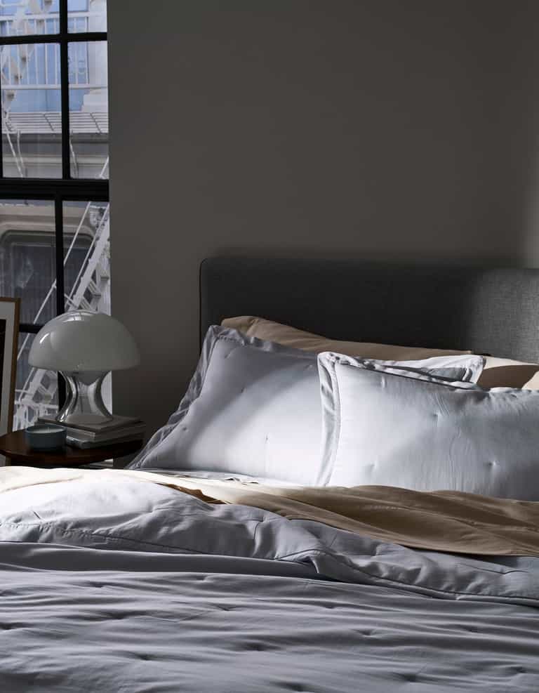 A bed with Cozy Earth Soft-Wash Cotton Quilted Shams in white and beige sits by a sunlit window. A bedside table holds a white lamp and books, while part of a gray upholstered headboard is seen.|Color:Shore