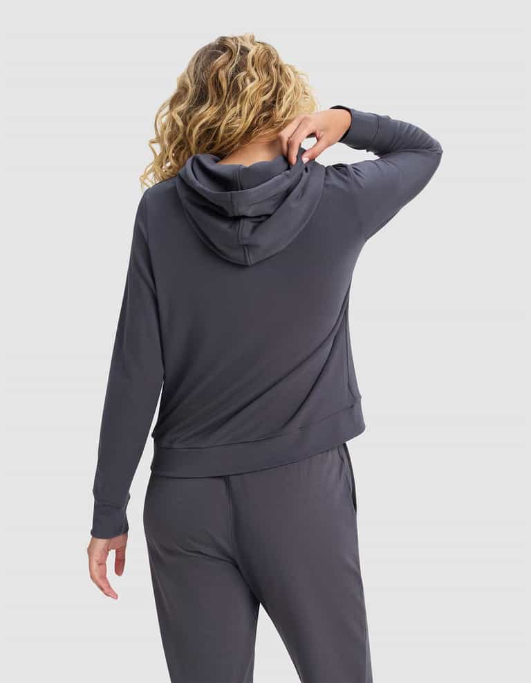 A woman with curly blonde hair, seen from the back, wears the Cozy Earth Women's Brushed Bamboo Hoodie in dark gray, paired with matching pants. She lifts one arm to touch her neck against a plain light gray background.|Color:Graphite