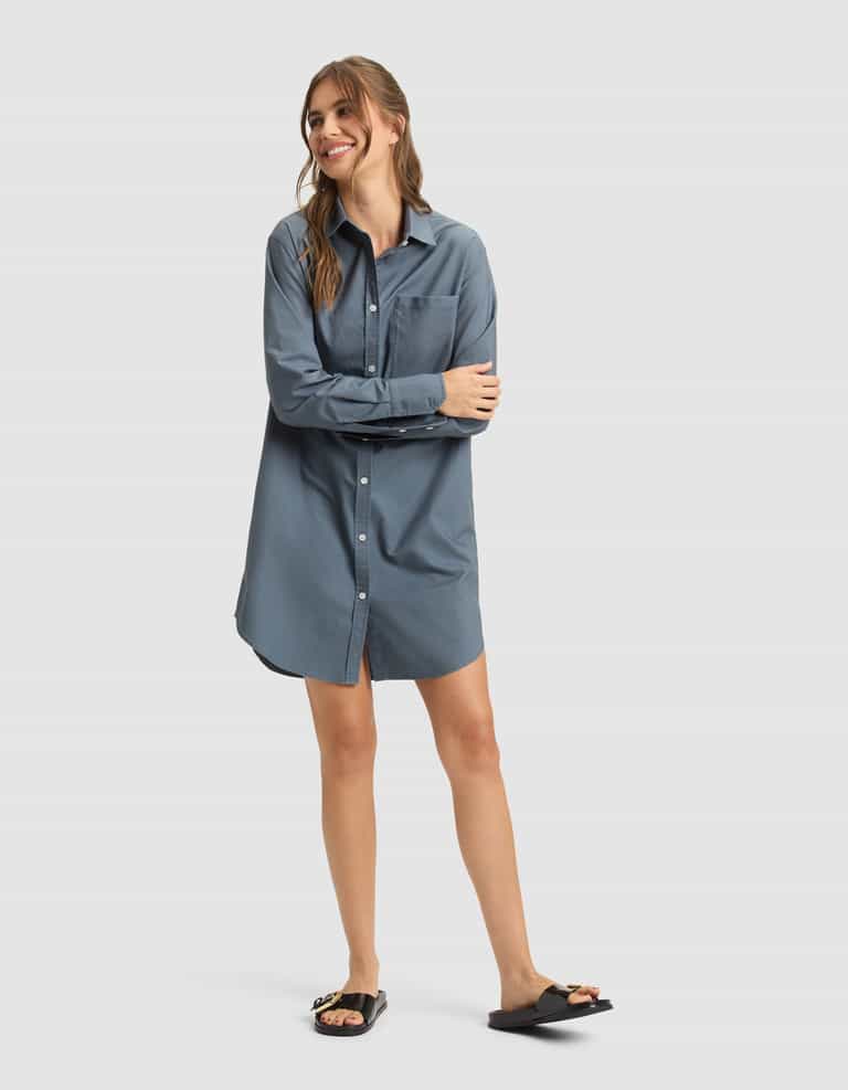A woman stands smiling with arms crossed, wearing the Cozy Earth Women's Camden Dress—a loose-fitting, long-sleeve blue shirt dress with front buttons—and black slide sandals. The background is a plain light gray. |Color:Dawn