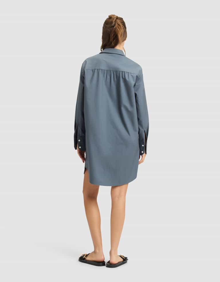 A woman with partially tied-up hair stands with her back to the camera, wearing the Cozy Earth Women's Camden Dress—a loose-fitting, long-sleeve blue shirt dress—and black flat sandals with chain details. The background is plain and light-colored. |Color:Dawn