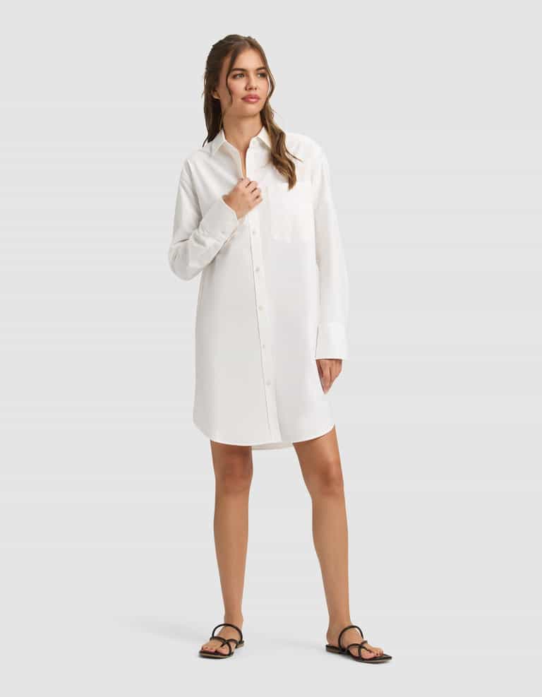 A woman with light brown hair stands against a plain background wearing the Cozy Earth Women's Camden Dress, a long-sleeved white button-up shirt dress with a front pocket, paired with black sandals. She gazes to the side with a relaxed expression. |Color:White