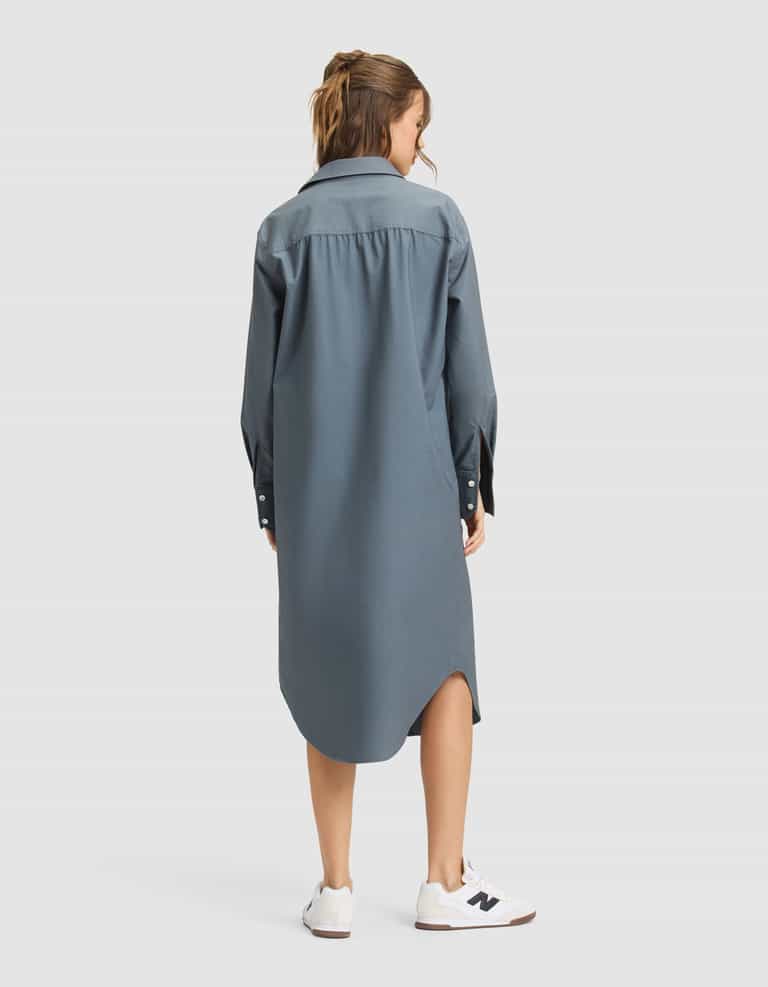 A woman faces away, wearing the Cozy Earth Women's Camden Midi Dress—a loose, long-sleeved blue button-down—paired with white sneakers on a plain light gray background. |Color:Dawn