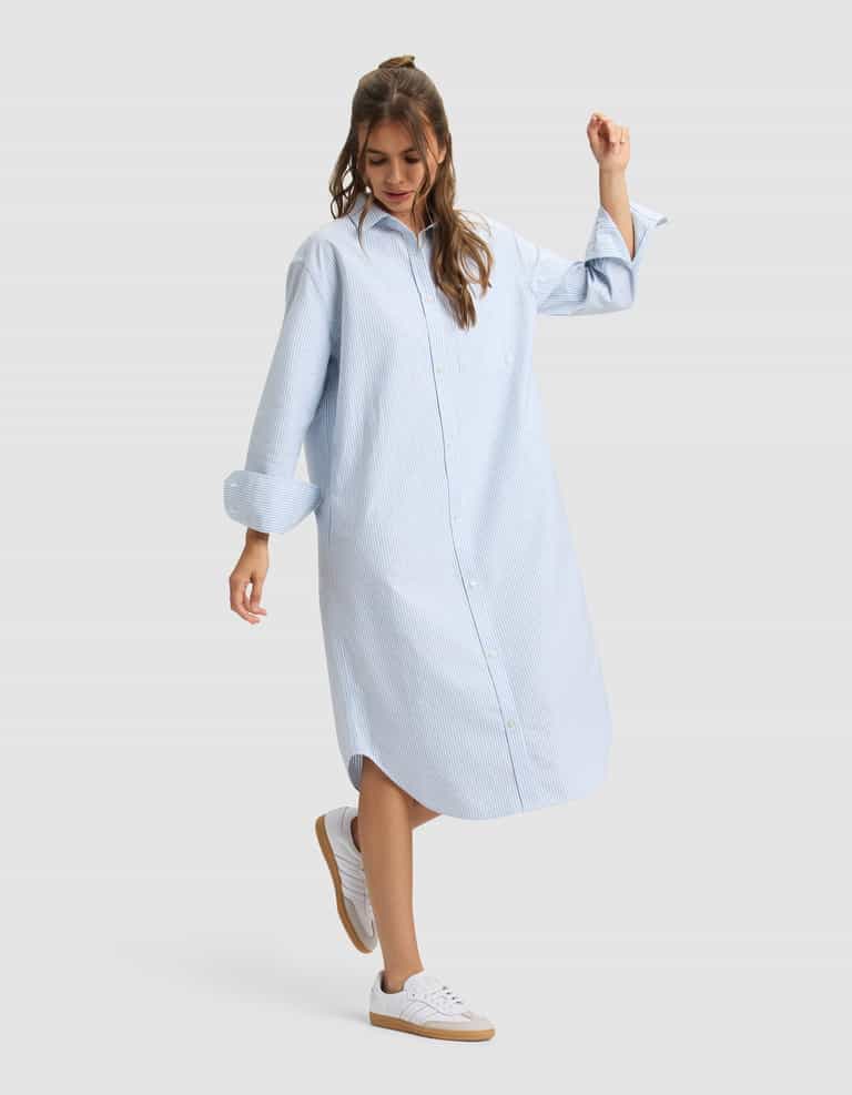 A woman wearing the Cozy Earth Women's Camden Midi Dress stands against a plain background, smiling and slightly lifting one foot. |Color:Blue Oxford Stripe