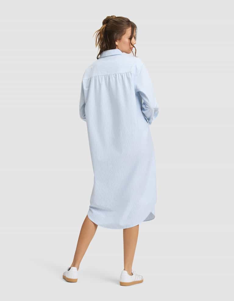 A woman faces away, wearing Cozy Earth's Women's Camden Midi Dress in light blue with subtle stripes and long sleeves, paired with white sneakers. Her hair is partially tied up, set against a plain light gray background. |Color:Blue Oxford Stripe
