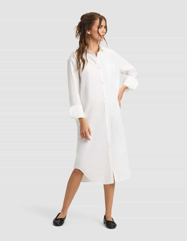 A woman wearing the Cozy Earth Women's Camden Midi Dress with rolled-up sleeves and black flats smiles, looking to the side with wavy brown hair styled half-up and one hand on her hip. |Color:White